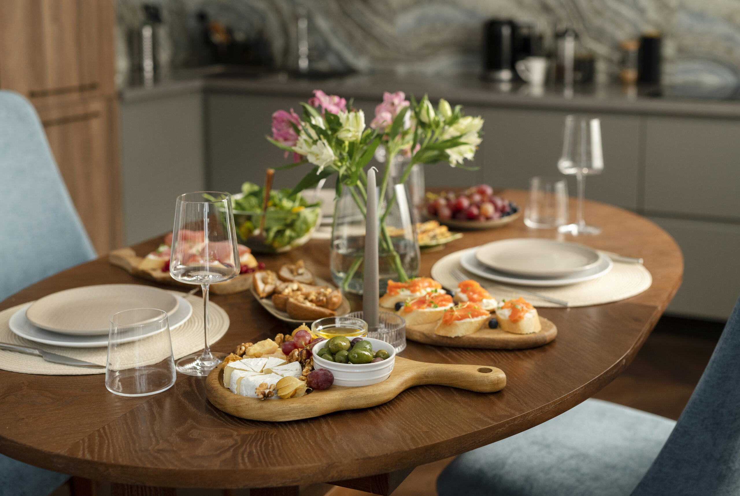 Wooden dining table set with flowers, candles, plates, and small food dishes in a modern kitchen.
